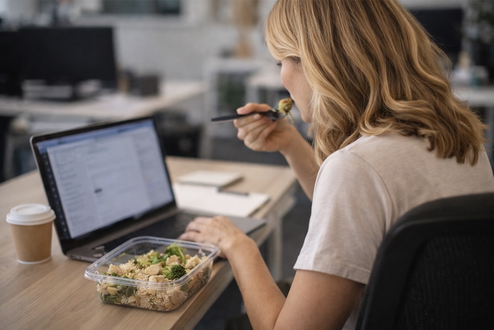 Frau isst nebenbei Mittagessen am Schreibtisch im Büro und arbeitet am Laptop, von hinten seitlich aufgenommen